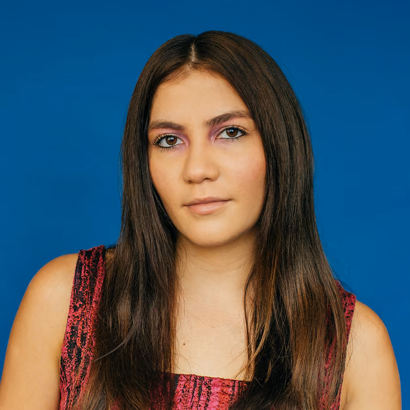 Woman with long brown hair wearing pink eyeshadow and a sleeveless pink patterned top on blue background.